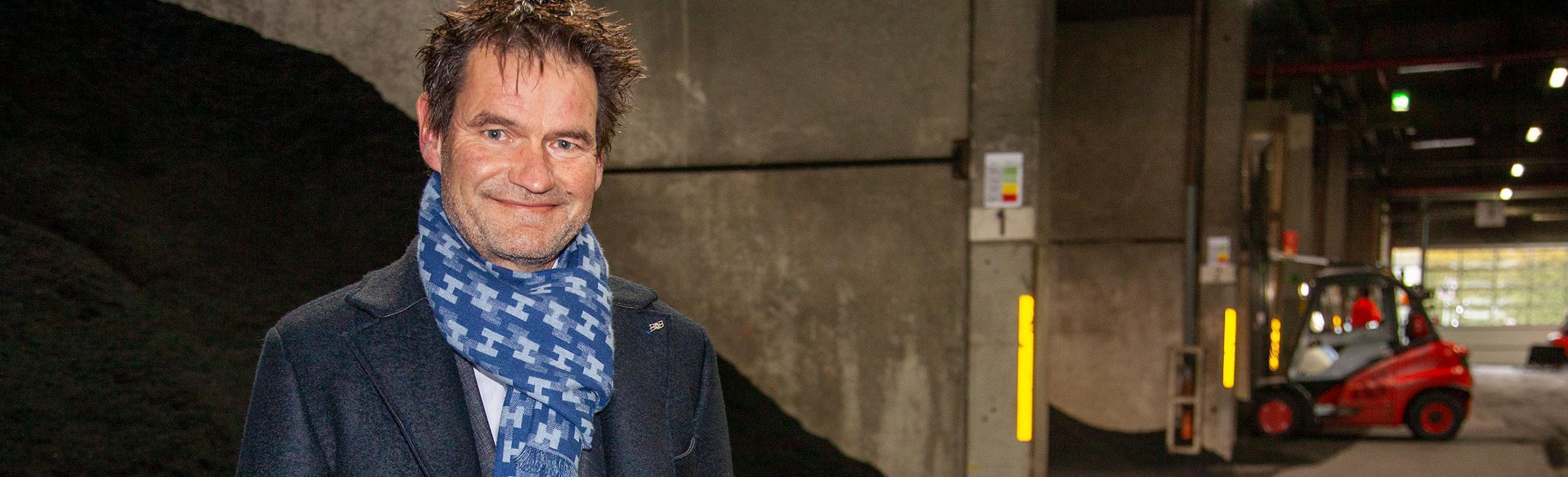 One of REGUPOL’s CEOs stands in an industrial hall wearing dark clothing and a blue patterned scarf. In the background are bulk materials and a red forklift in operation.