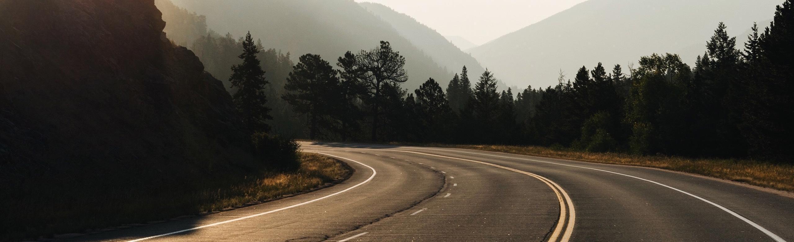 A winding rural road runs through a forested mountain landscape. The road is empty and curves between trees and rocky hills, with sunlight creating a hazy, atmospheric mood.