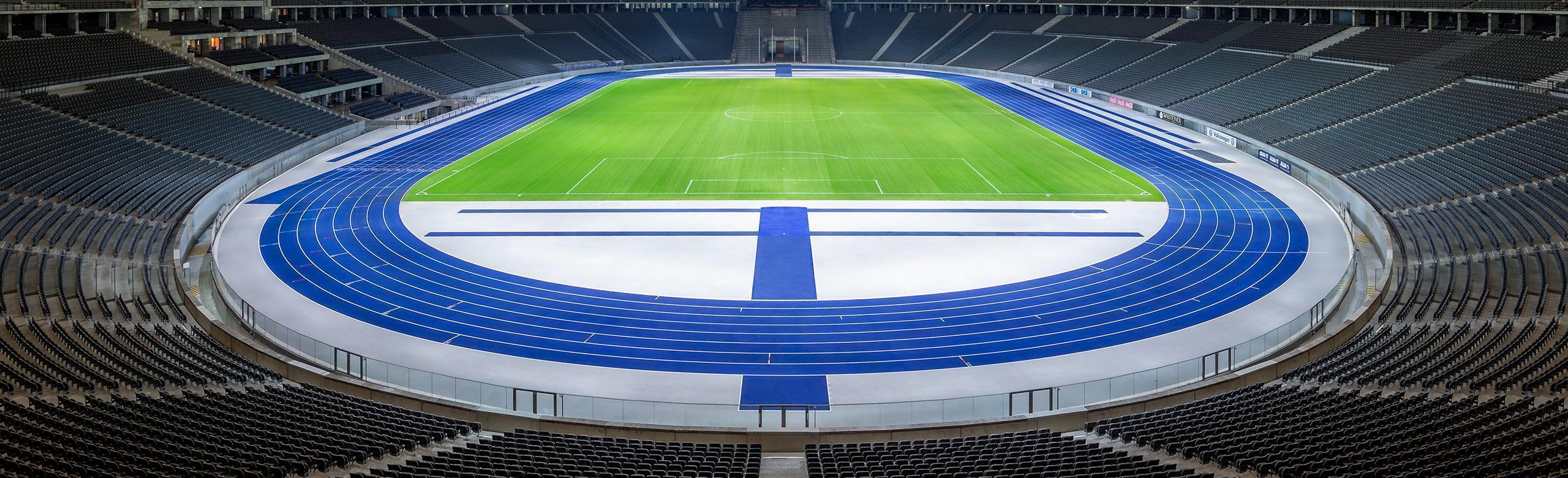 View into the empty Olympic Stadium Berlin featuring the distinctive blue REGUPOL running track around the green field. The grandstands are empty